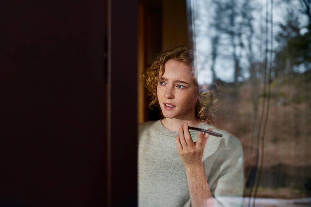En kvinna håller i en mobil och tittar ut genom fönstret.