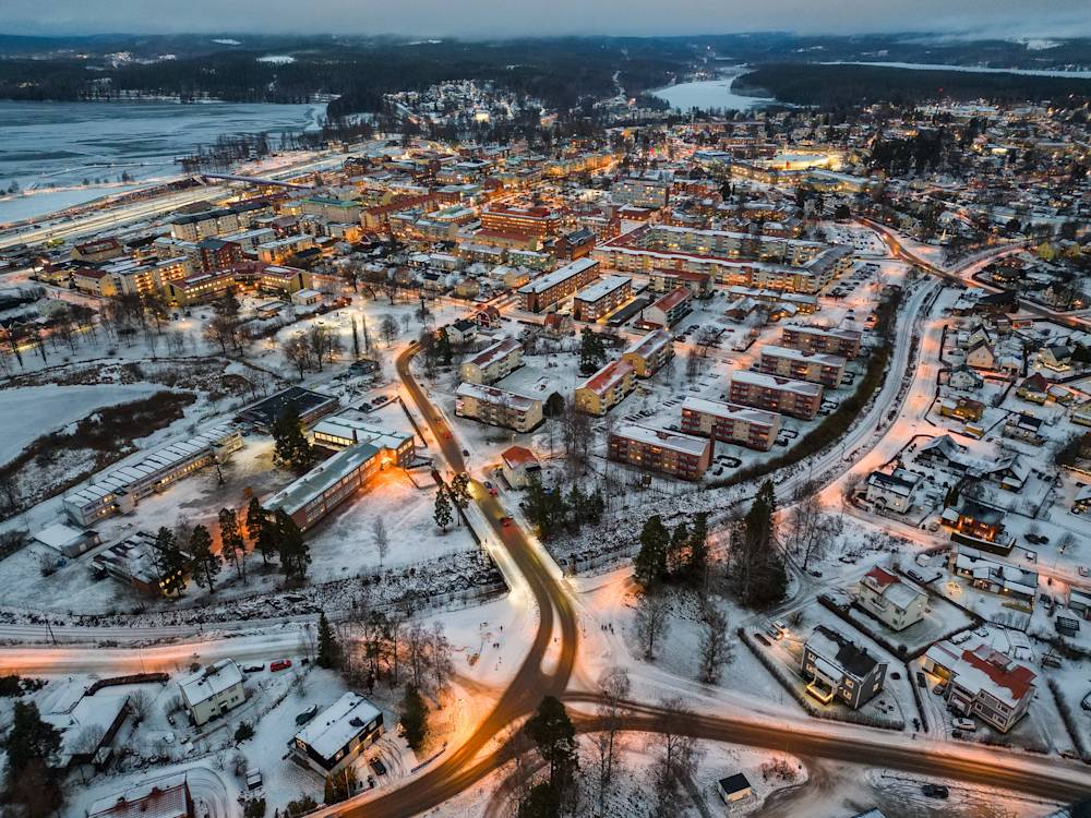 Översiktsbild av ett samhälle under de mörka timmarna av vinterhalvåret. Byggnaderna är upplysta och på gatorna mellan husen syns vägar täckta av snö.