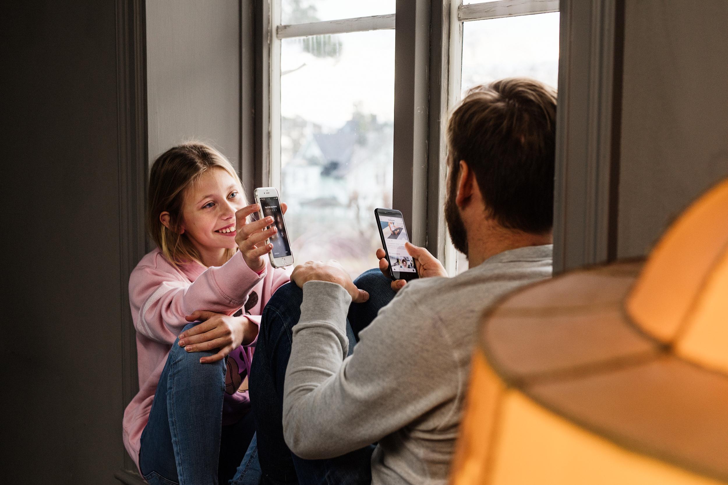 En flicka och pappa sitter vid ett fönster och tittar på sina mobiler. Flickan visar sin mobil för pappan. 