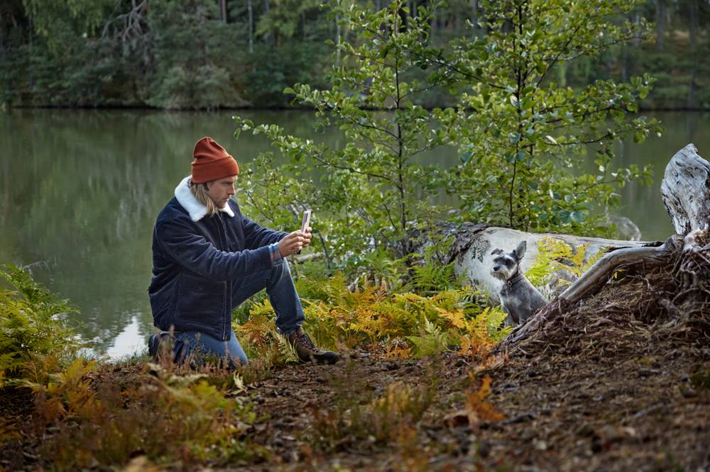 Man fotograferar med mobilen när han är ute i höstlik natur med en sjö i bakgrunden tillsammans med sin hund 