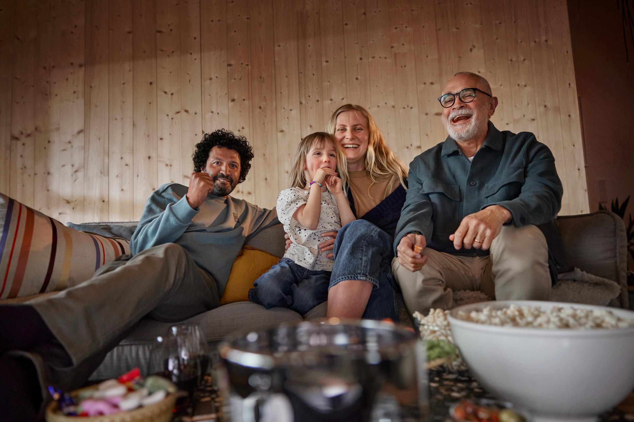 Familj med tre generationer tillsammans sitter i soffan och tittar på tv och har kul tillsammans - en stor popcornskål syns i förgrunden. 