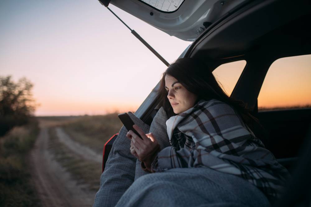 En tjej sitter i bilen en sensommarkväll, omvirad i en filt och tittar på sin mobil med solnedgången bakom sig. 