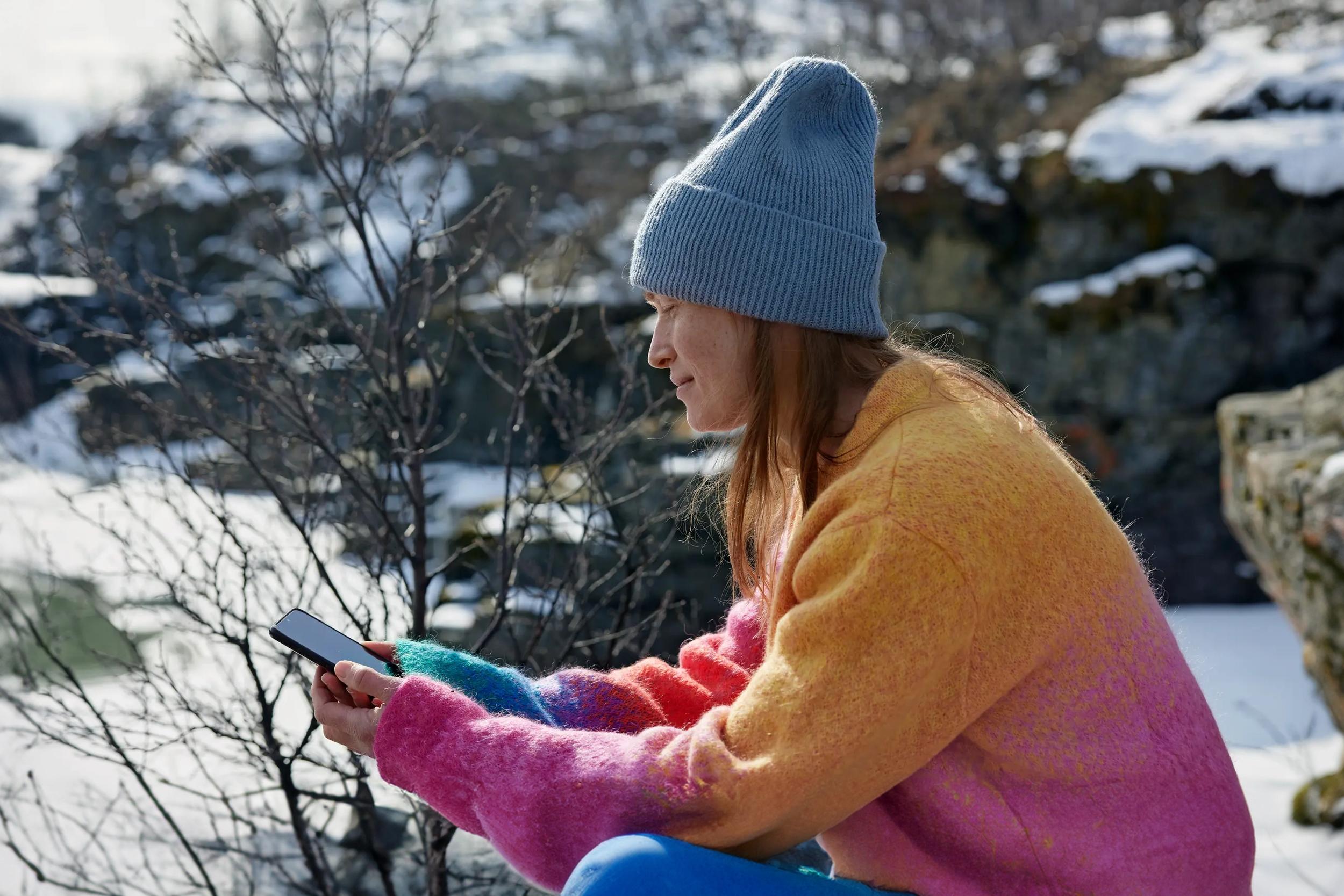 Kvinna som sitter på fjället och surfar på sin mobil. I bakgrunden syns snö och skog.